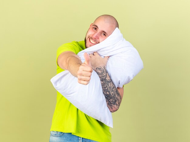 Souriant jeune homme malade tient oreiller et pouces vers le haut isolé sur mur vert olive