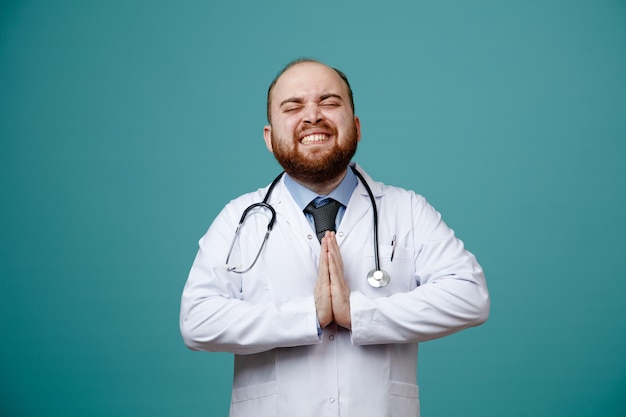 Souriant jeune homme médecin portant un manteau médical et un stéthoscope autour du cou faisant un geste de namaste avec les yeux fermés isolés sur fond bleu