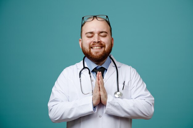 Souriant jeune homme médecin portant un manteau médical et un stéthoscope autour du cou et des lunettes sur la tête montrant le geste de namaste avec les yeux fermés isolés sur fond bleu