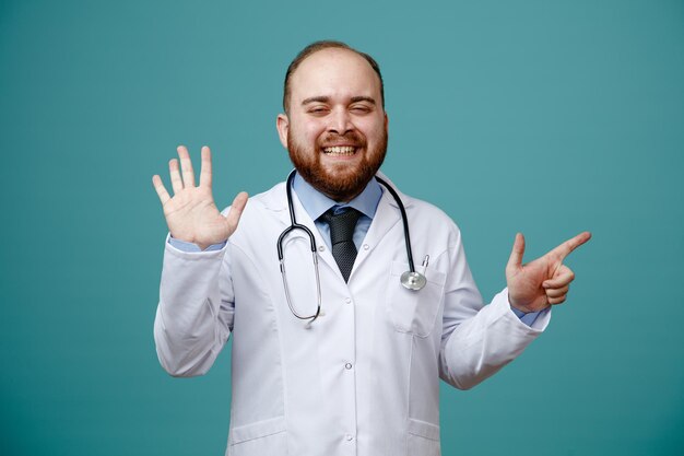 Souriant jeune homme médecin portant un manteau médical et un stéthoscope autour du cou regardant la caméra montrant cinq avec la main pointant vers le côté isolé sur fond bleu