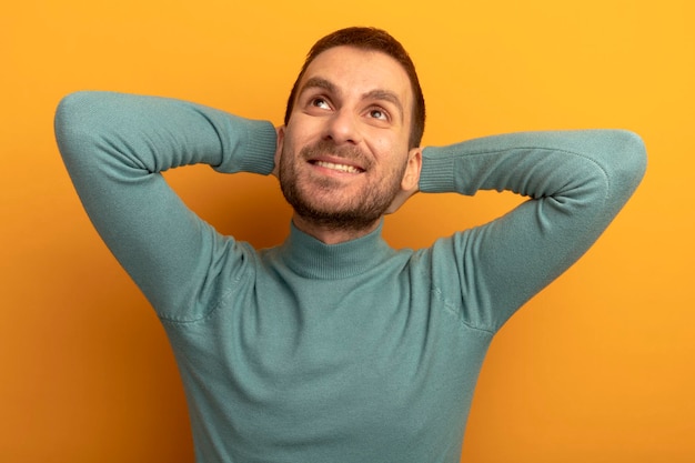 Souriant jeune homme mettant les mains derrière la tête en levant isolé sur mur orange