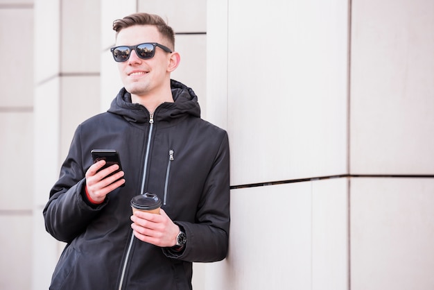 Souriant jeune homme avec mobile dans la main tenant une tasse de café à emporter