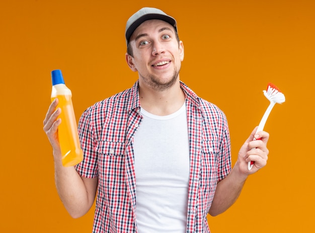 Souriant jeune homme nettoyant portant une casquette tenant un agent de nettoyage avec une brosse isolée sur fond orange