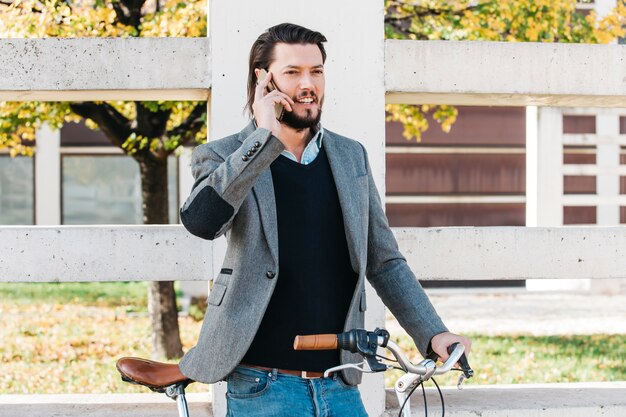 Souriant jeune homme parlant au téléphone portable debout avec vélo dans le parc