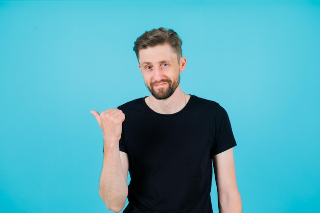 Souriant jeune homme pointe vers la gauche avec le pouce sur fond bleu