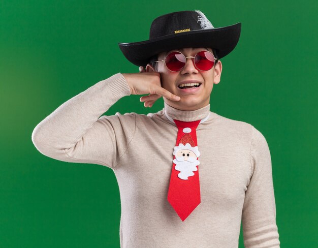 Souriant jeune homme portant chapeau avec cravate de Noël et lunettes montrant le geste d'appel téléphonique isolé sur le mur vert