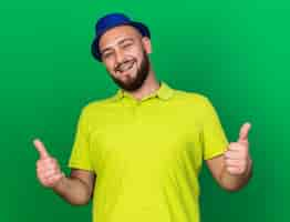 Photo gratuite souriant jeune homme portant un chapeau de fête bleu montrant les pouces vers le haut