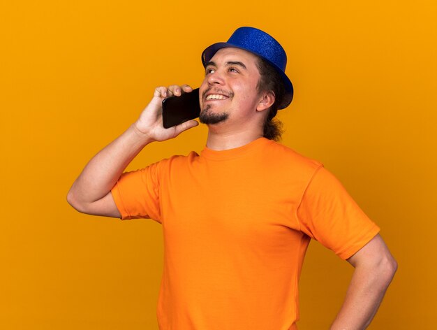 Souriant jeune homme portant un chapeau de fête parle au téléphone en mettant la main sur la hanche