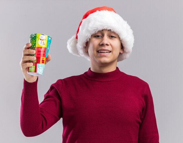 Souriant jeune homme portant un chapeau de Noël tenant une tasse de Noël isolé sur un mur blanc