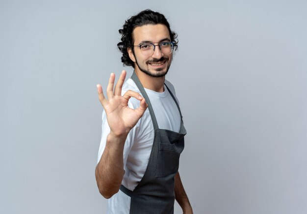 Souriant jeune homme de race blanche barbier portant des lunettes et bande de cheveux ondulés en uniforme debout en vue de profil faisant signe ok