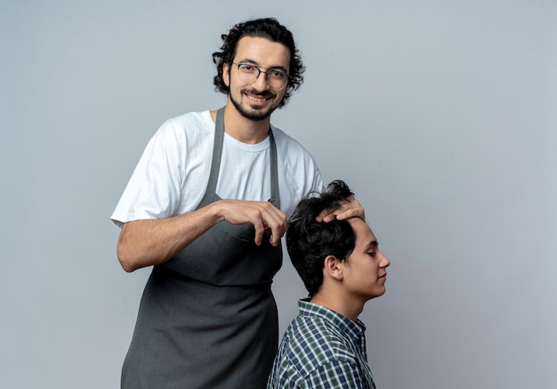 Souriant jeune homme de race blanche coiffeur portant des lunettes et bande de cheveux ondulés en uniforme faisant la coupe de cheveux pour son jeune client isolé sur fond blanc avec espace de copie