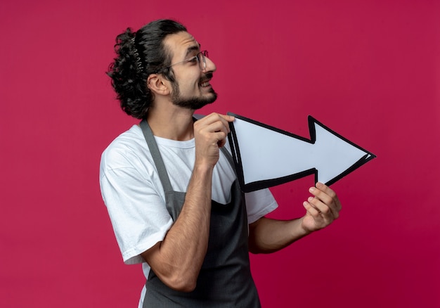 Souriant jeune homme de race blanche coiffeur portant des lunettes et bande de cheveux ondulés en uniforme tenant la flèche qui pointe sur le côté et regardant côté isolé sur fond cramoisi