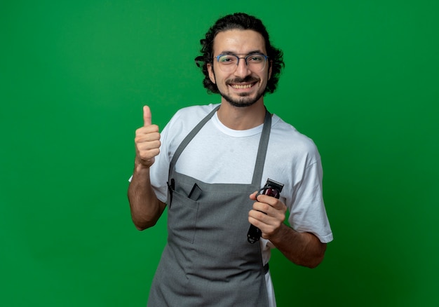 Souriant jeune homme de race blanche coiffeur portant des lunettes et bande de cheveux ondulés en uniforme tenant une tondeuse à cheveux et montrant le pouce vers le haut isolé sur fond vert avec espace copie