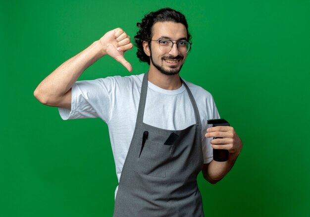 Souriant jeune homme de race blanche coiffeur portant des lunettes et bande de cheveux ondulés en uniforme tenant un vaporisateur montrant le pouce vers le bas isolé sur fond vert avec espace copie