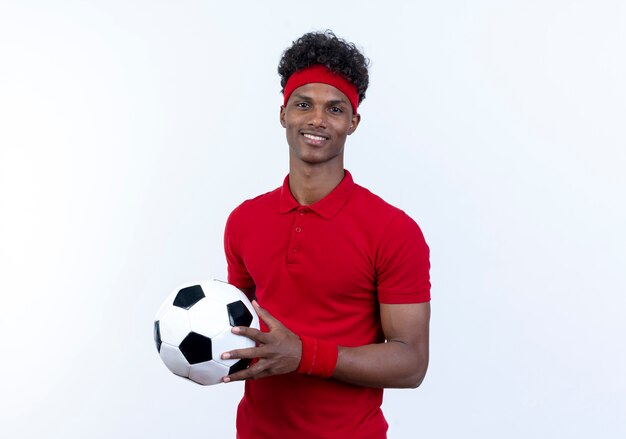 Souriant jeune homme sportif afro-américain portant bandeau et bracelet tenant ballon isolé sur blanc