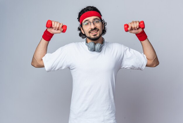 Souriant jeune homme sportif portant un bandeau avec un bracelet et des écouteurs sur le cou faisant de l'exercice avec des haltères