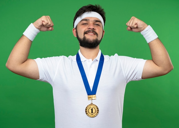 Photo gratuite souriant jeune homme sportif portant bandeau et bracelet avec médaille montrant un geste fort isolé sur vert