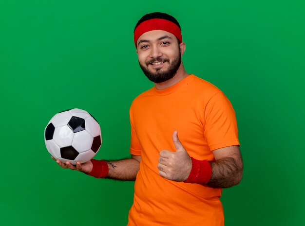 Souriant jeune homme sportif portant bandeau et bracelet tenant la balle sur le côté et montrant le pouce vers le haut isolé sur fond vert