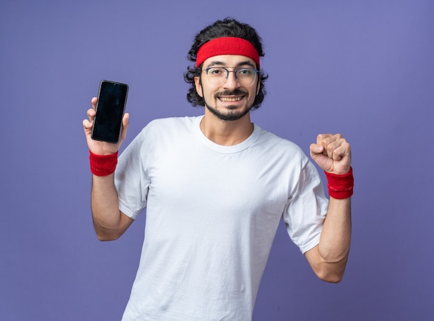 Souriant jeune homme sportif portant un bandeau avec un bracelet tenant un téléphone montrant un geste oui