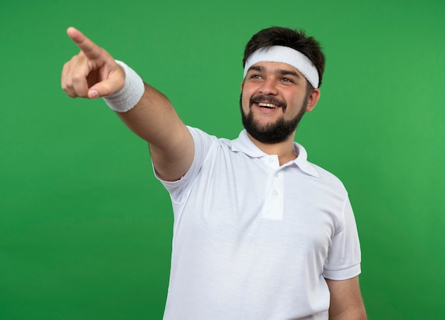 Souriant jeune homme sportif regardant côté portant des points de bandeau et de bracelet sur le côté isolé sur mur vert