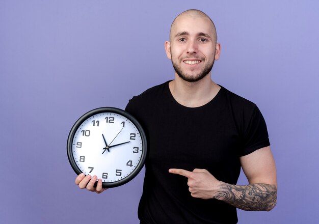 Souriant jeune homme sportif tenant et points à l'horloge murale isolée sur violet