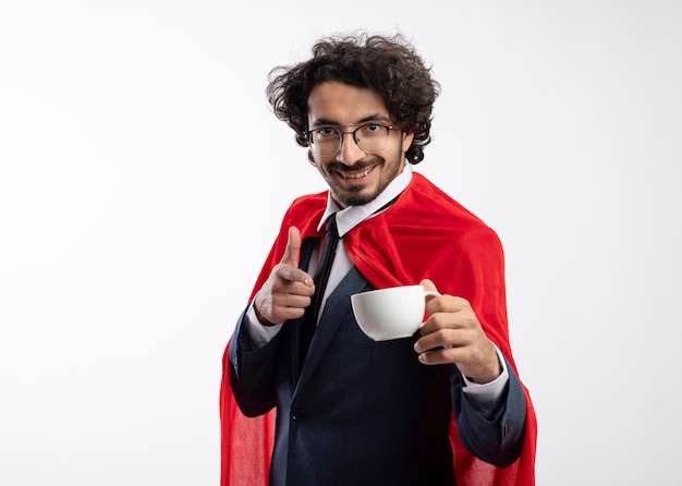 Souriant jeune homme de super-héros caucasien dans des lunettes optiques portant un costume avec une cape rouge tient et pointe vers la tasse