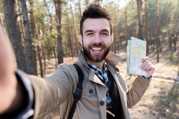 Souriant jeune homme tenant la carte dans la main prenant selfie dans la forêt