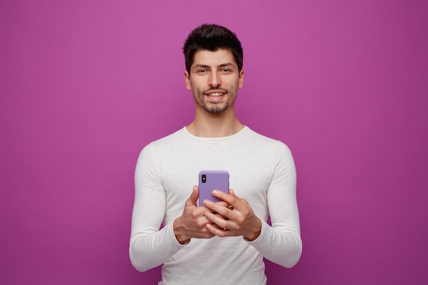 Souriant jeune homme tenant un téléphone portable regardant la caméra isolée sur fond violet