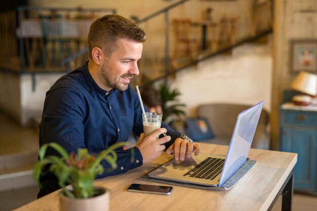 Souriant jeune homme travaillant sur l'ordinateur dans un café-bar restaurant confortable