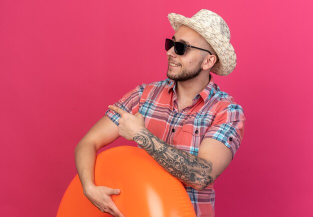 Souriant jeune homme voyageur caucasien avec chapeau de plage de paille dans des lunettes de soleil tenant un anneau de bain regardant et pointant sur le côté isolé sur fond rose avec espace de copie