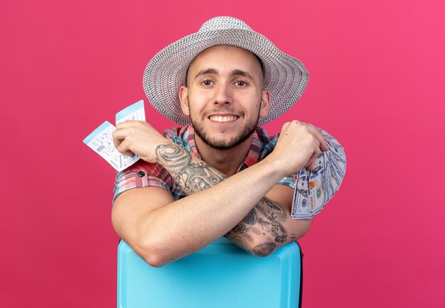 Souriant jeune homme voyageur caucasien avec chapeau de plage de paille tenant des billets d'avion et de l'argent debout derrière une valise isolé sur fond rose avec espace de copie