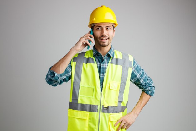 Souriant jeune ingénieur masculin portant un casque de sécurité et un uniforme regardant le côté en gardant la main sur la taille tout en parlant au téléphone isolé sur fond blanc