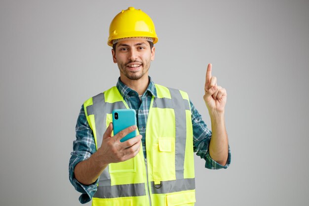 Souriant jeune ingénieur masculin portant un casque de sécurité et un uniforme tenant un téléphone portable regardant la caméra pointant vers le haut isolé sur fond blanc