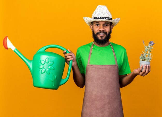 Souriant jeune jardinier afro-américain portant un chapeau de jardinage tenant un arrosoir avec une fleur dans un pot de fleurs