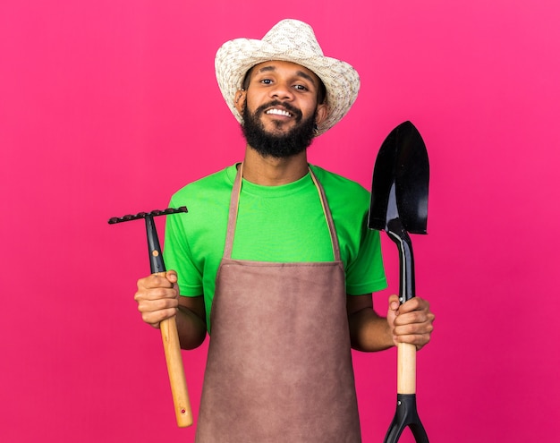 Souriant jeune jardinier afro-américain portant un chapeau de jardinage tenant une pelle avec un râteau isolé sur un mur rose