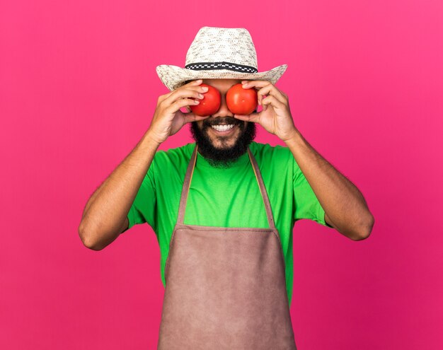 Souriant jeune jardinier afro-américain portant un chapeau de jardinage tenant une tomate et montrant un geste de regard isolé sur un mur rose