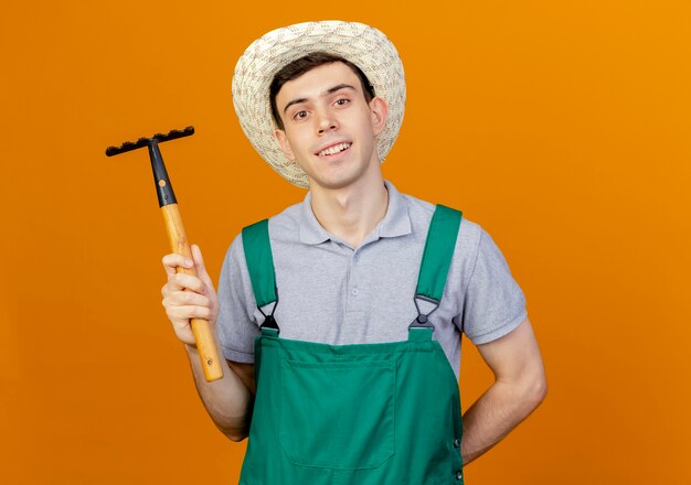 Souriant jeune jardinier mâle portant chapeau de jardinage détient râteau