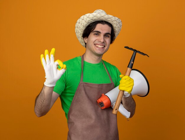 Souriant jeune jardinier mâle en uniforme portant chapeau de jardinage avec des gants tenant haut-parleur avec râteau montrant oui geste isolé sur orange