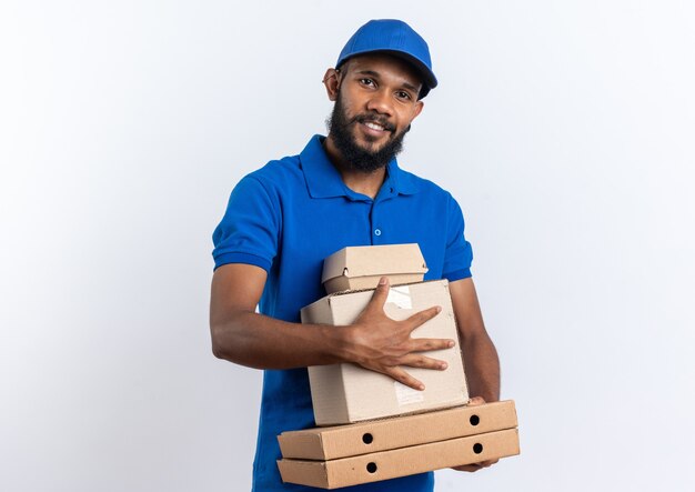 Souriant jeune livreur afro-américain tenant une boîte en carton et un emballage de nourriture sur des boîtes à pizza isolées sur un mur blanc avec espace de copie