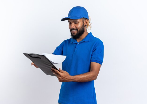 Souriant jeune livreur afro-américain tenant et regardant le presse-papiers isolé sur un mur blanc avec espace de copie