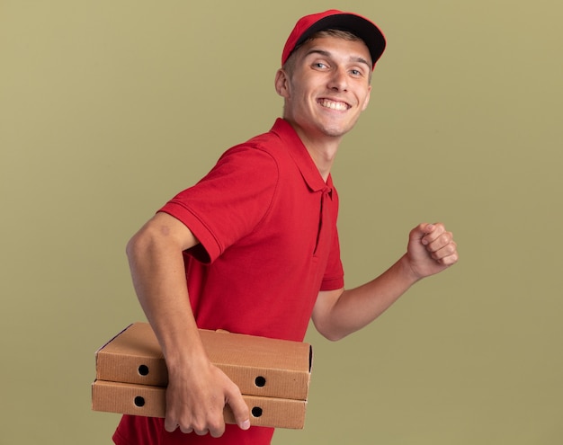 Souriant jeune livreur blond se tient sur le côté tenant des boîtes à pizza faisant semblant de courir isolé sur un mur vert olive avec espace de copie