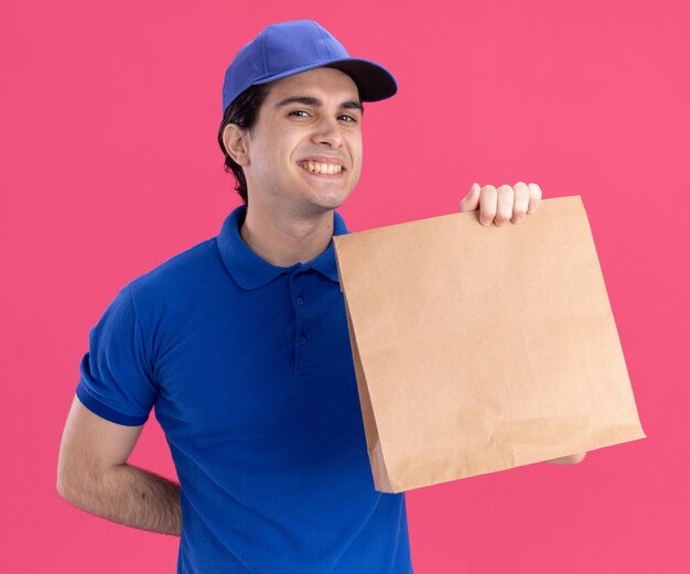 Souriant jeune livreur caucasien en uniforme bleu et casquette tenant un paquet de papier gardant la main derrière le dos isolé sur un mur rose