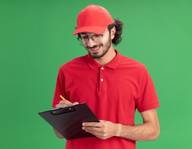 Souriant jeune livreur caucasien en uniforme rouge et casquette portant des lunettes écrivant sur le presse-papiers avec un crayon