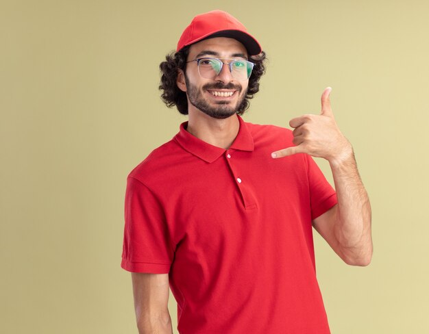 Souriant jeune livreur caucasien en uniforme rouge et casquette portant des lunettes faisant un geste lâche isolé sur un mur vert olive avec espace de copie