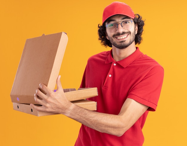 Souriant Jeune Livreur Caucasien En Uniforme Rouge Et Casquette Portant Des Lunettes Tenant Des Paquets De Pizza En Ouvrant Un