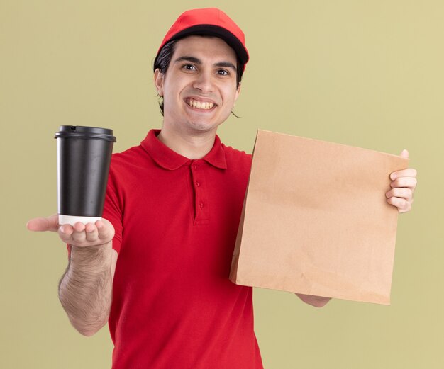 souriant jeune livreur caucasien en uniforme rouge et casquette tenant un paquet de papier et étirant une tasse de café en plastique vers la caméra regardant la caméra isolée sur fond vert olive