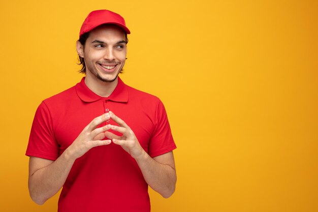 souriant jeune livreur portant l'uniforme et la casquette gardant les mains ensemble regardant le côté isolé sur fond jaune avec espace de copie