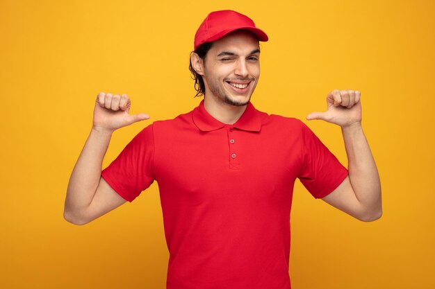 souriant jeune livreur portant l'uniforme et la casquette regardant la caméra clignant de l'œil pointant vers lui-même isolé sur fond jaune