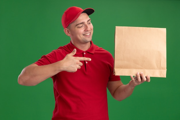 Photo gratuite souriant jeune livreur portant l'uniforme et la casquette tenant et points paquet alimentaire papier isolé sur mur vert