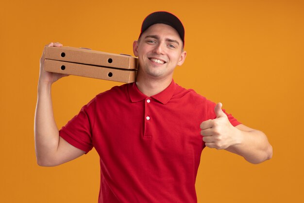 Souriant jeune livreur en uniforme avec capuchon tenant des boîtes de pizza sur l'épaule montrant le pouce vers le haut isolé sur un mur orange